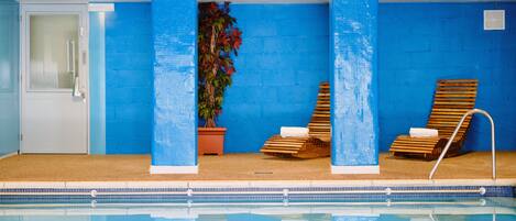 A swimming pool with blue walls and columns, two wooden loungers, and a potted plant.