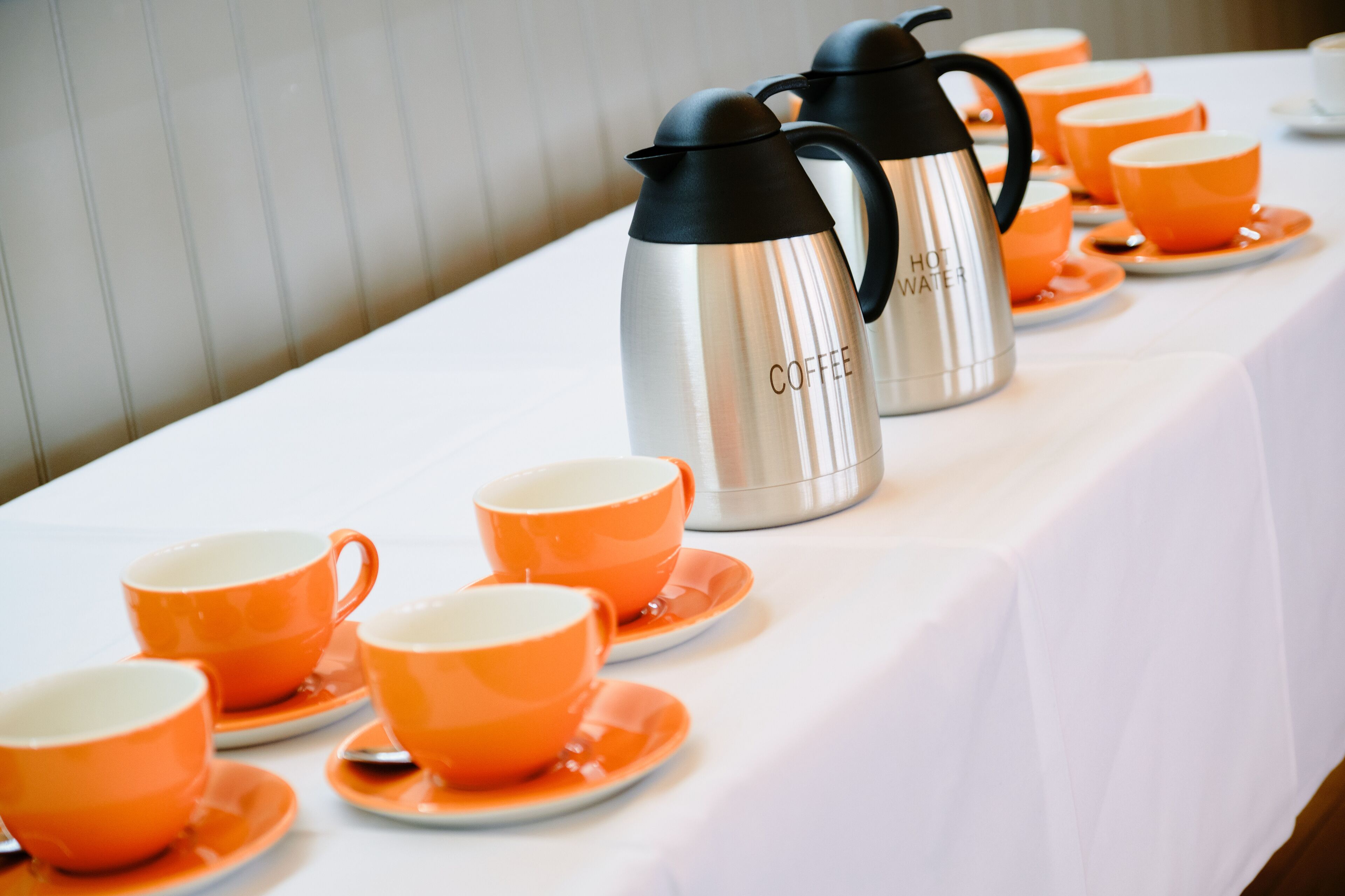 A table set with orange cups and saucers, and two stainless steel hot water carafes labeled "HOT WATER" and "COFFEE".