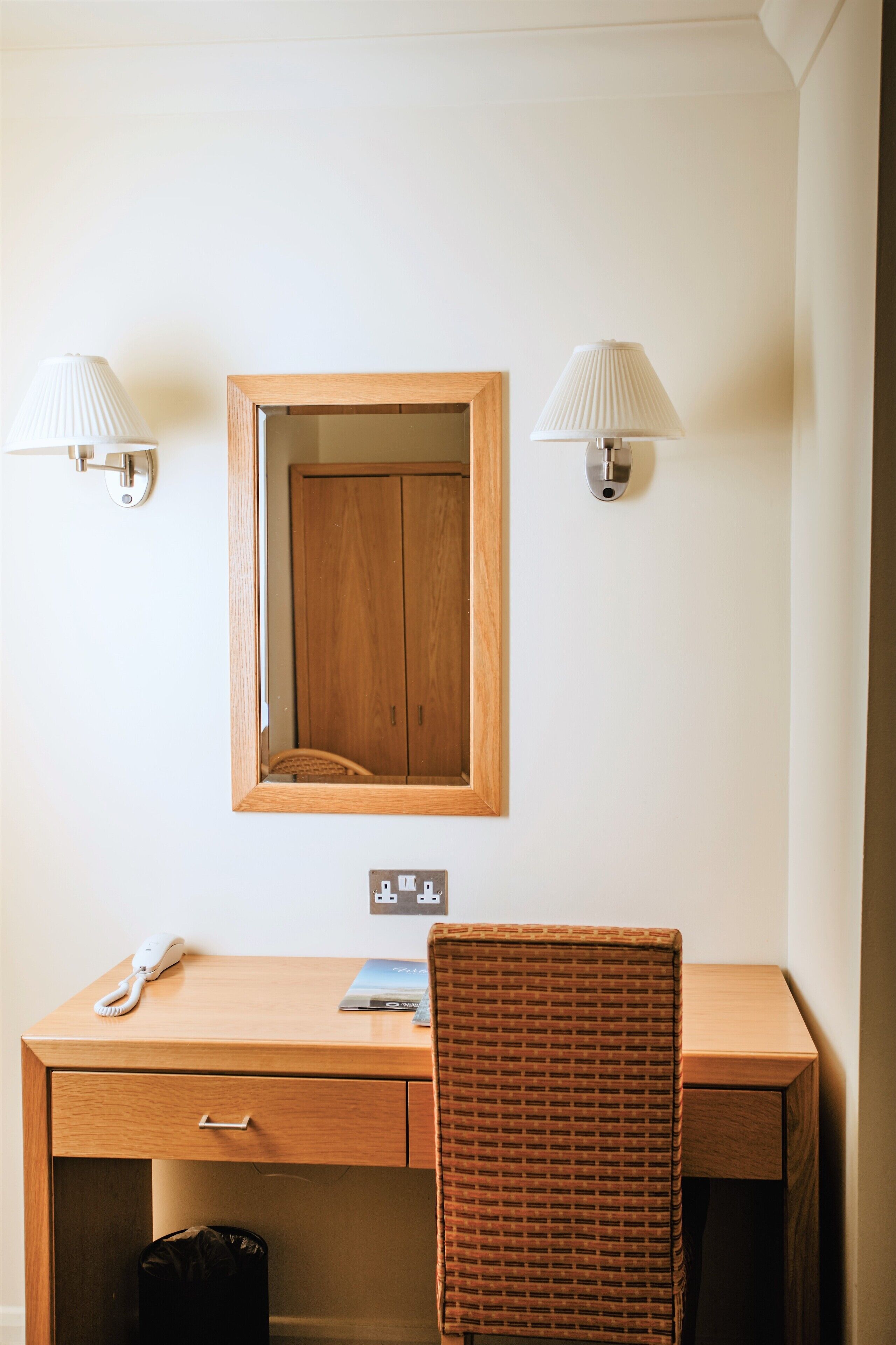 Une chambre d’hôtel avec un bureau en bois, une chaise en osier, un miroir et deux lampes fixées au mur.