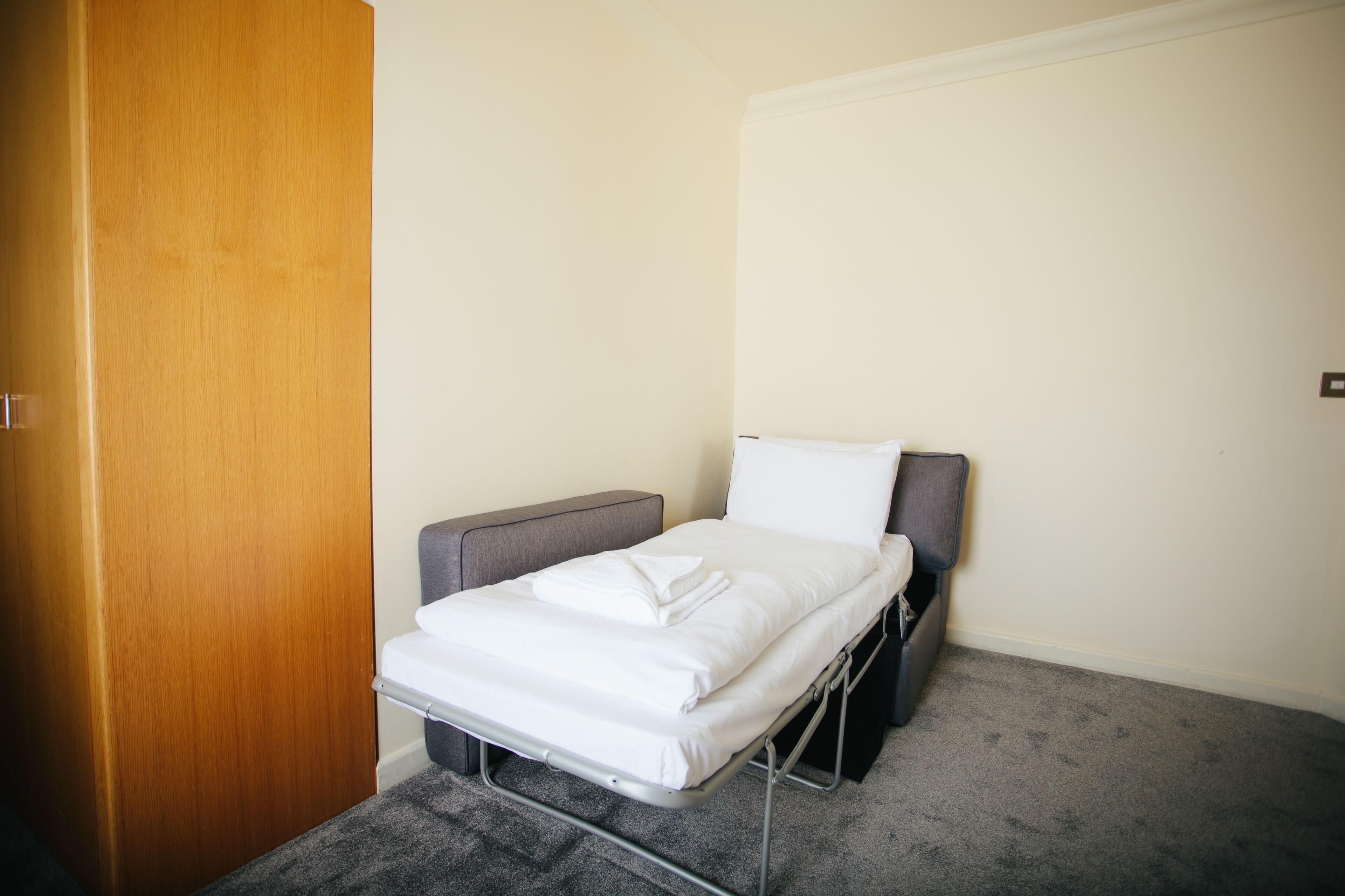 Une petite chambre avec un lit simple, une porte en bois, une tête de lit grise et un matelas blanc.