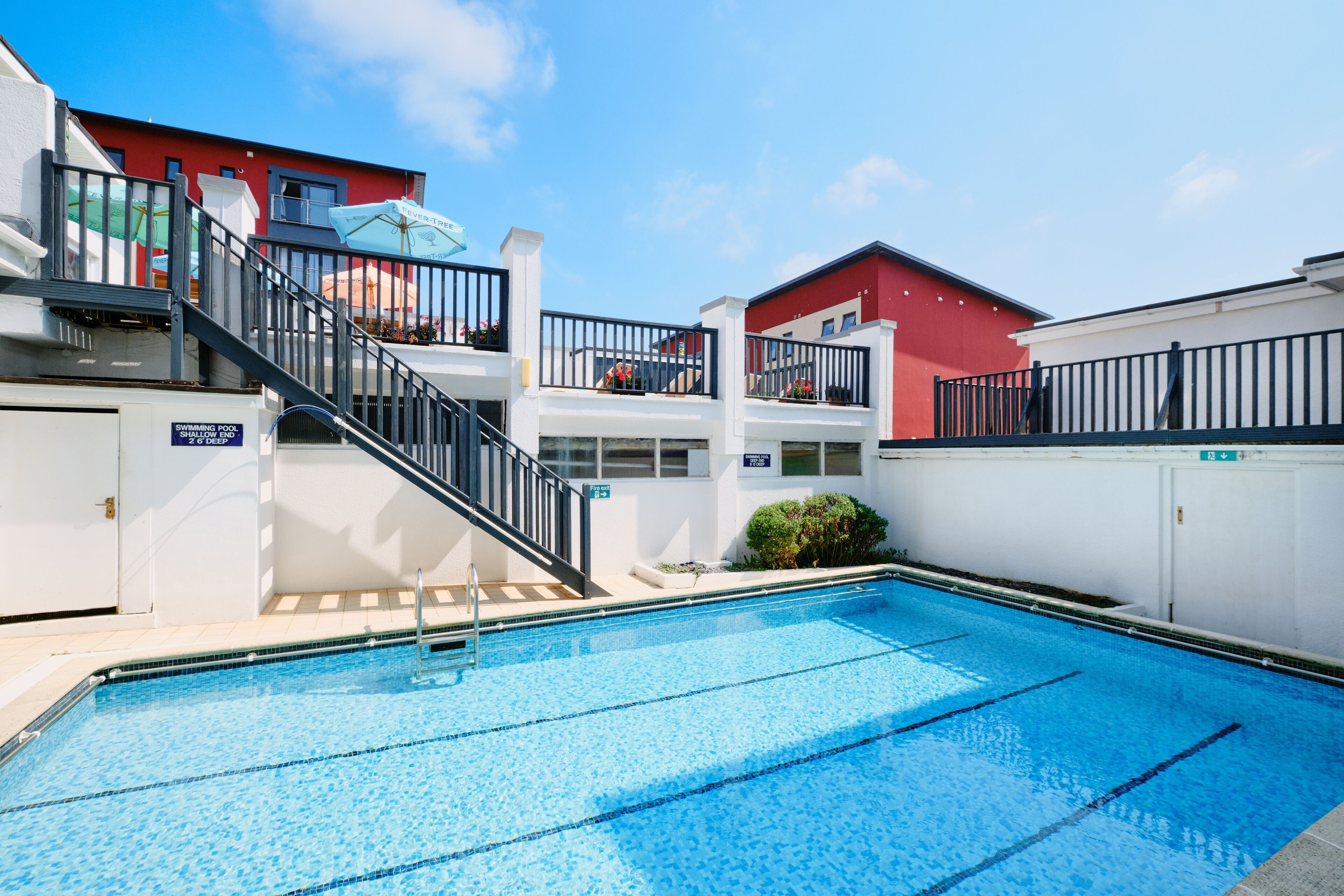 A swimming pool with a clear blue water, surrounded by white walls and a red building in the background.