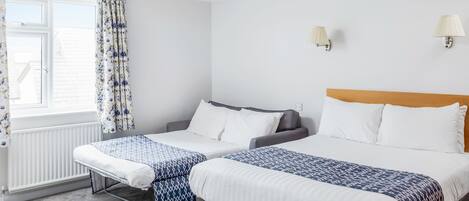 A hotel room with two beds, a window with curtains, a radiator, and wall-mounted lamps.
