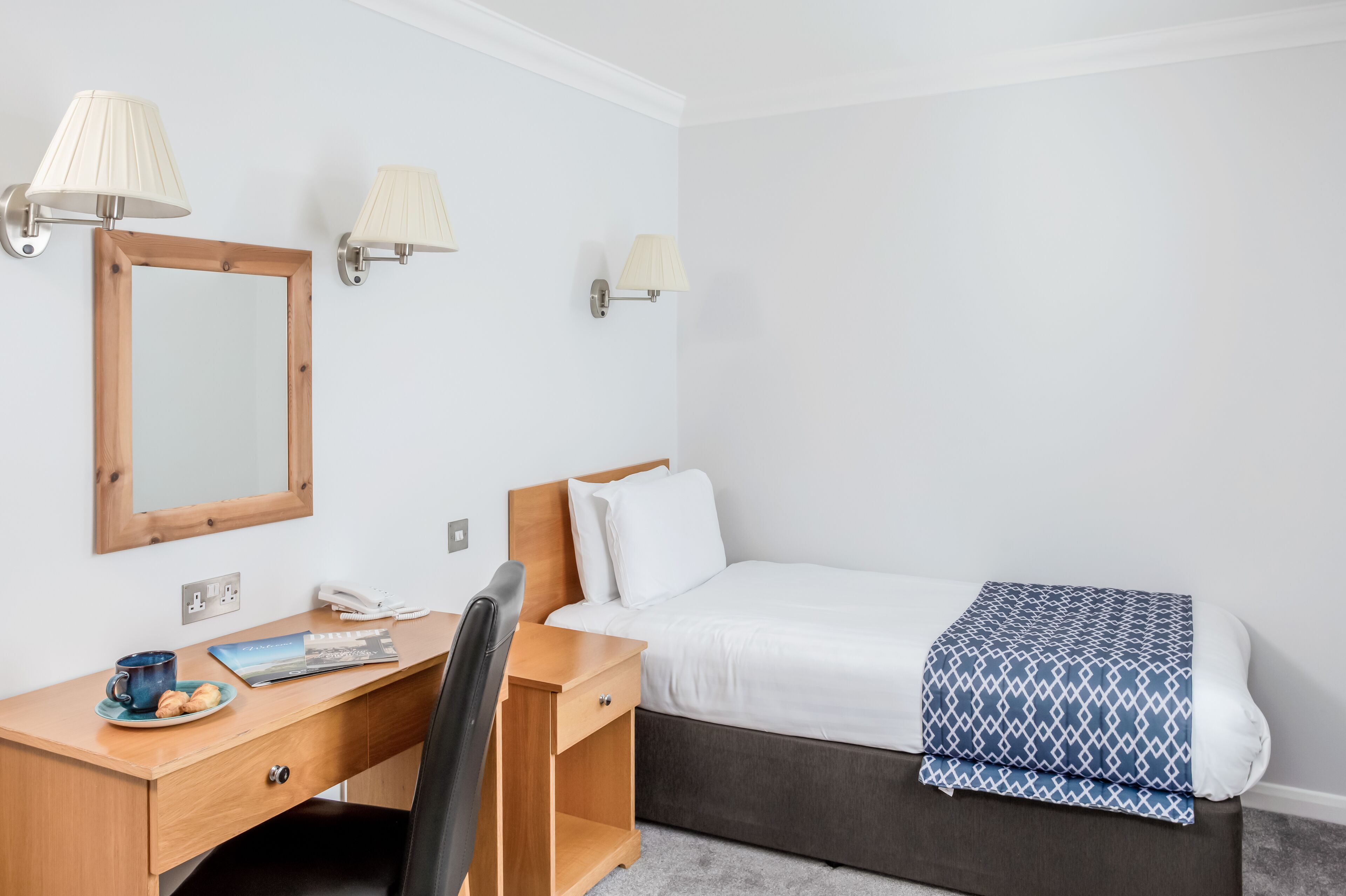 A hotel room with a wooden desk, a bed with a blue patterned blanket, and two wall-mounted lamps.