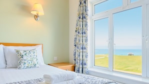A hotel room with a bed, a nightstand, a lamp, a window with a view of the sea, and a curtain with a floral pattern. - Kilbirnie Hotel (Newquay)