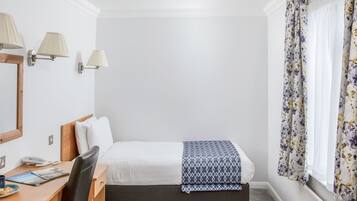 A hotel room with a bed, a desk with a chair, a telephone, and a window with floral curtains.