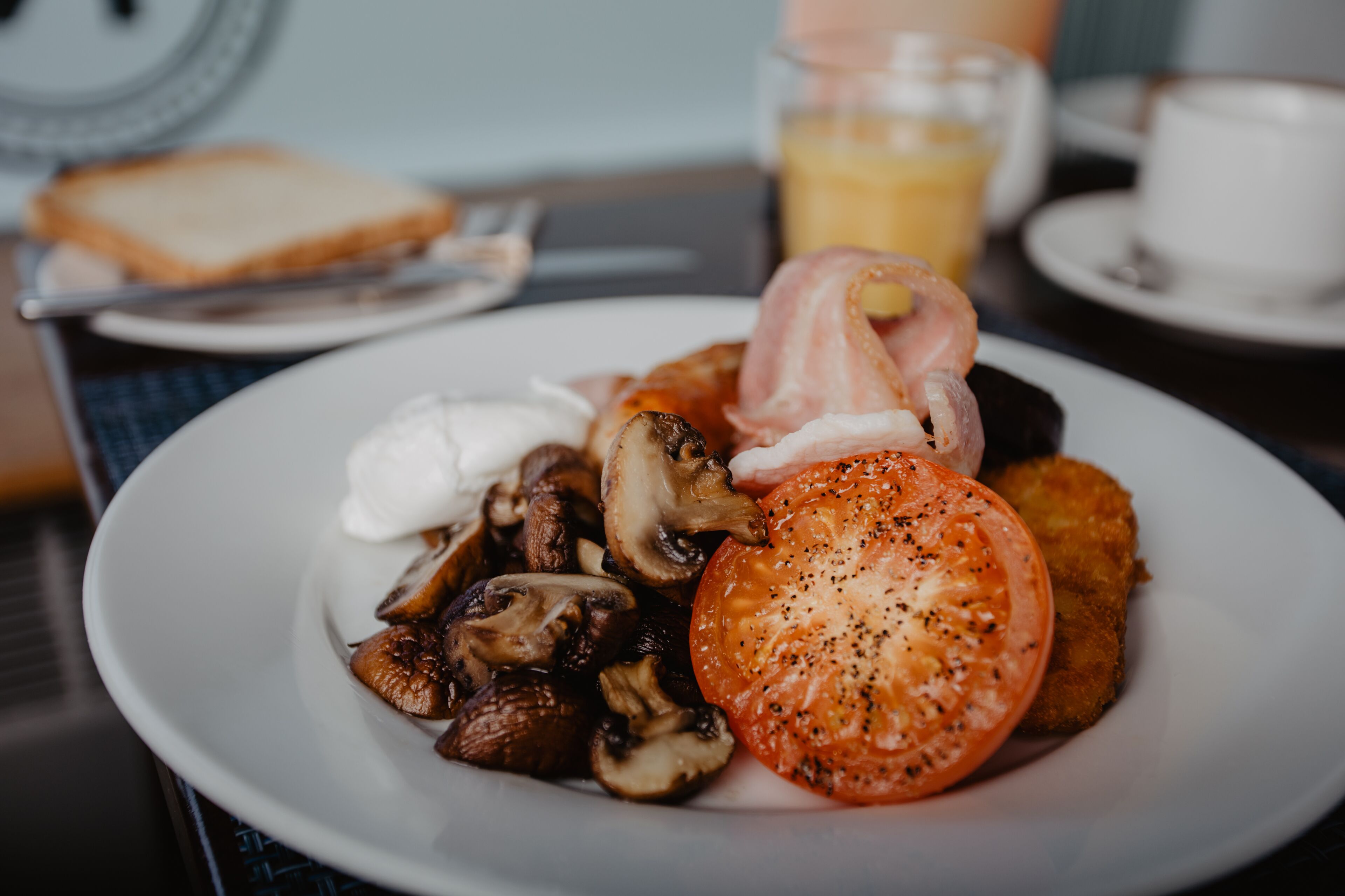 A plate of breakfast food including mushrooms, a tomato, and a fried item, with toast and a cup of coffee in the background.