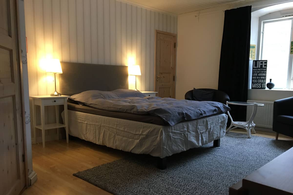 Un dormitorio con cama, dos lámparas de noche, una silla, una mesita y un ventanal con cortinas.