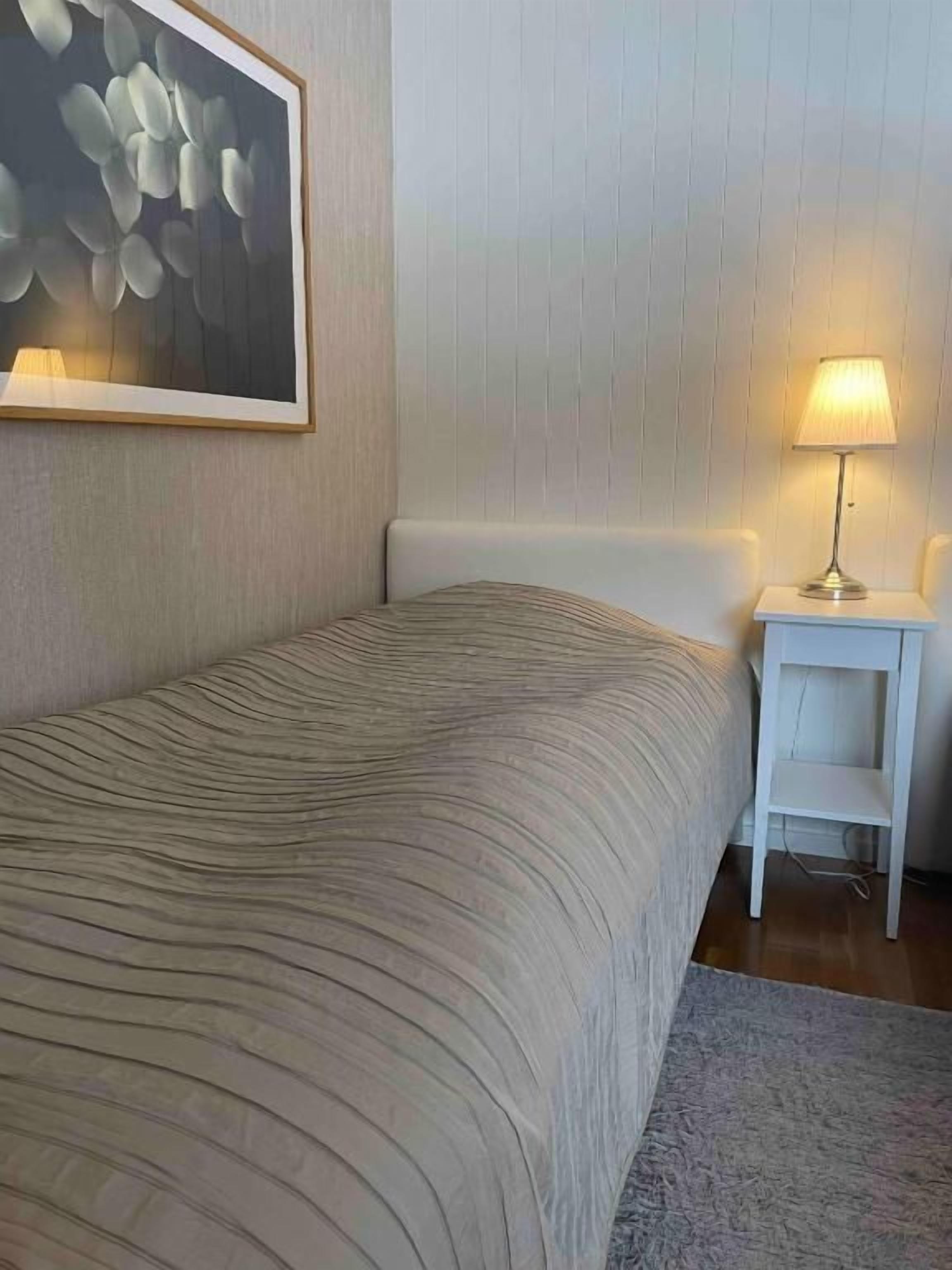 Un dormitorio con cama, mesita de noche, lámpara y un cuadro en la pared.