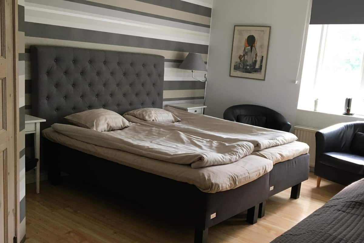 Un dormitorio con cabecera capitonada, cama con ropa de cama color beige, escritorio con lámpara, una silla y ventana con vistas.