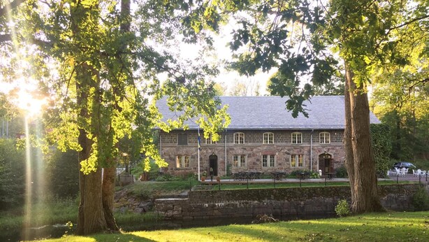 Un bâtiment en pierre à l’architecture traditionnelle, entouré d’arbres et d’une zone herbeuse.