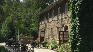 Une terrasse extérieure avec des tables et des chaises, un bâtiment en pierre doté de fenêtres en arc de cercle et un jardin agrémenté de fleurs et de verdure.
