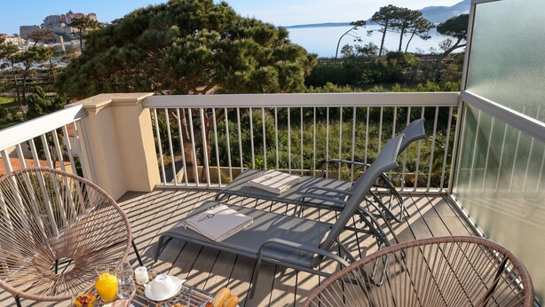 Un balcon avec une table dressée pour le petit-déjeuner, deux chaises en osier et une vue sur la mer et les arbres.