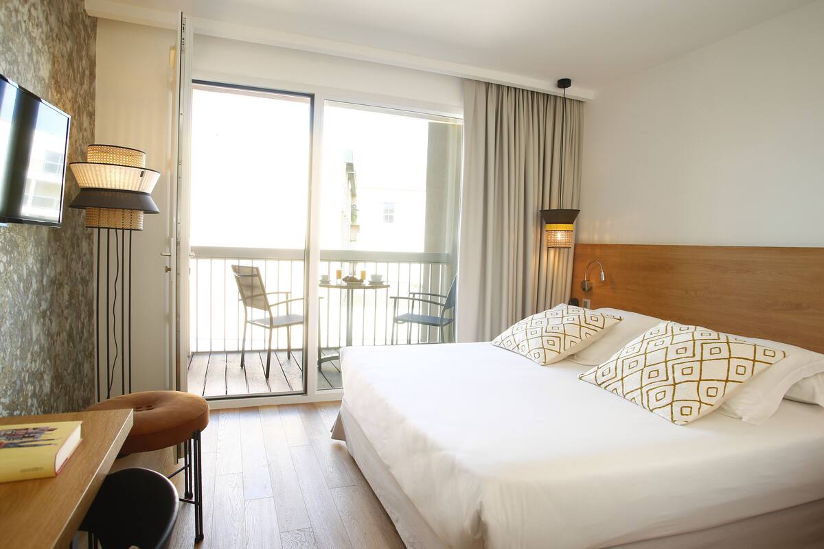 Habitación de hotel con cama, escritorio, silla, televisor y balcón con sillas.
