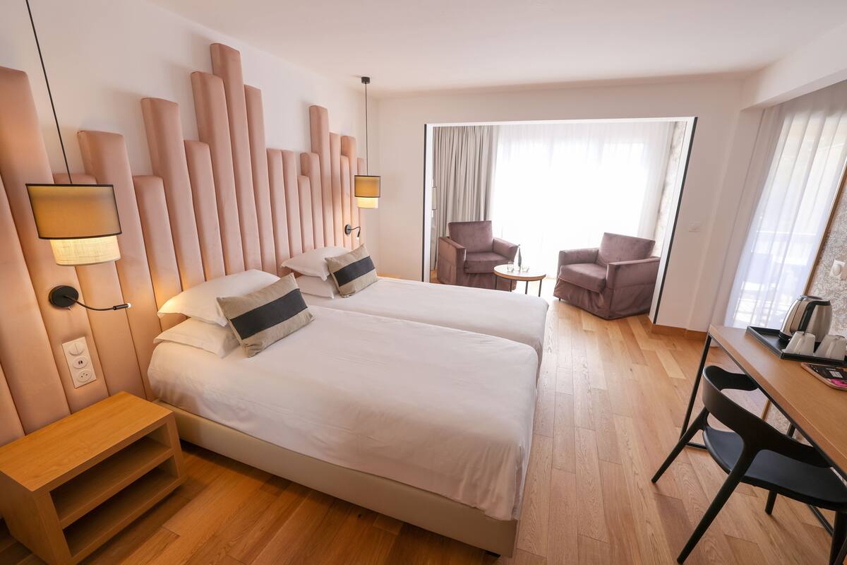 Habitación de hotel con dos camas, un escritorio y una silla. Hay una ventana con cortinas y vistas al exterior.