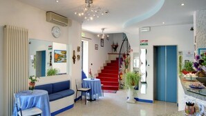 A hotel lobby with a blue sofa, a round table, a staircase, and a reception area. - Hotel Acquazzurra (Rimini)