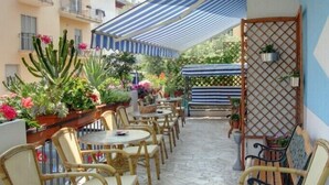 An outdoor patio area with round tables and wicker chairs, surrounded by potted plants and a striped awning. - Hotel Acquazzurra (Rimini)