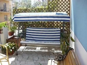 Un espace extérieur ensoleillé, aménagé avec des sièges, un banc à rayures et un auvent, entouré de plantes en pot et d’une clôture en treillis.