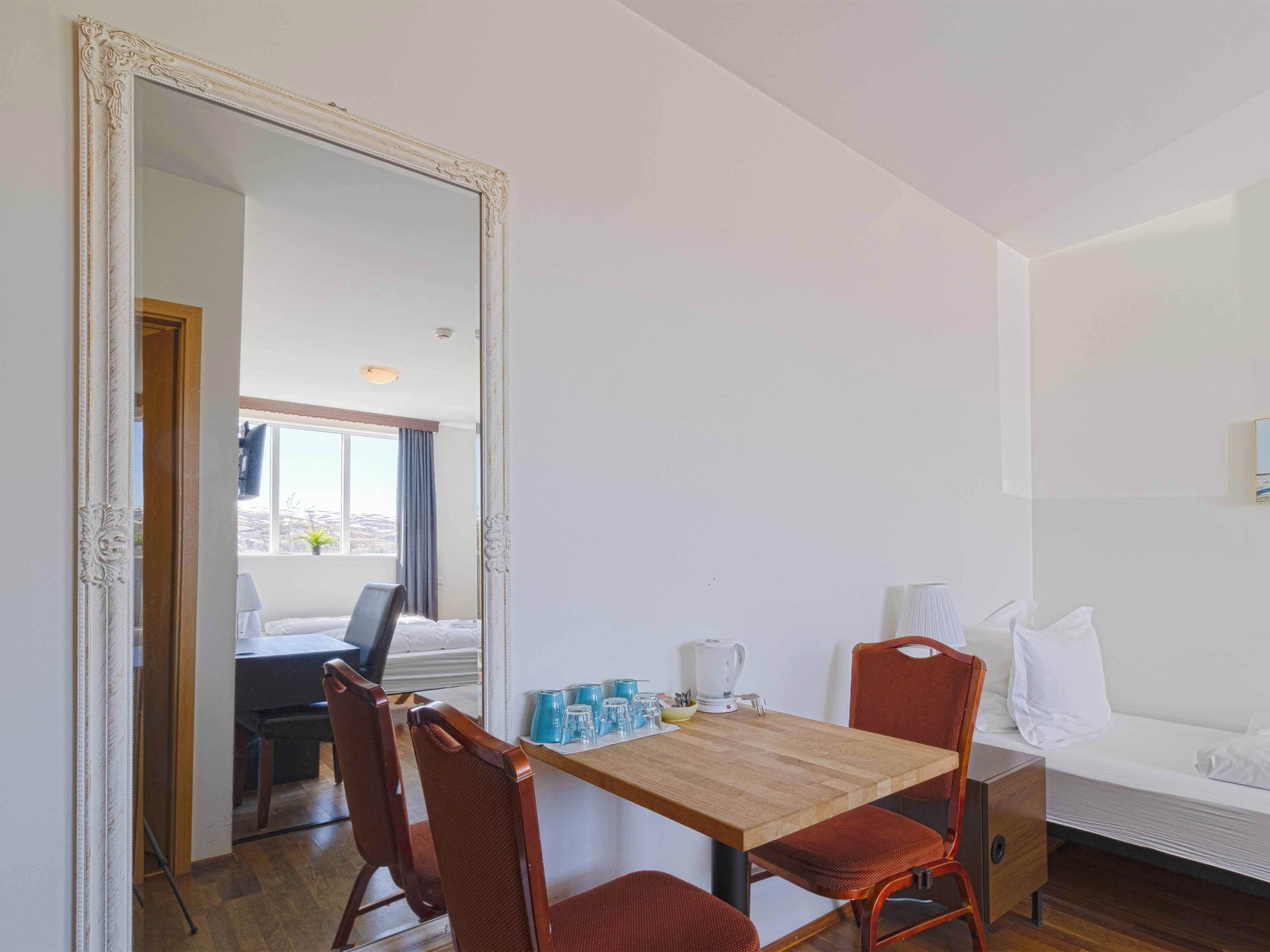 A hotel room with a wooden dining table, two red chairs, a white sofa, and a large mirror.