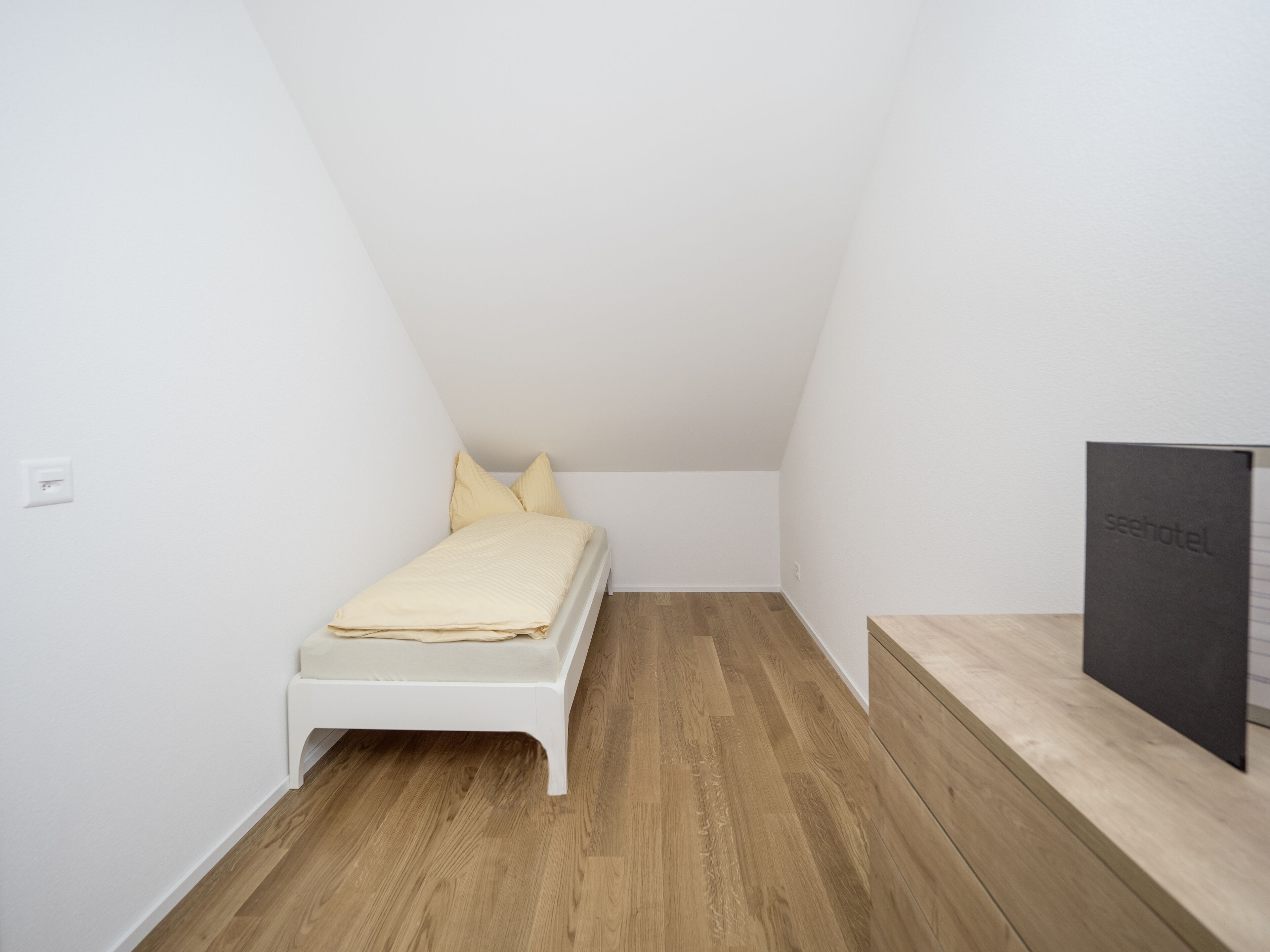 Une petite chambre blanche avec un lit simple, un plafond en pente et un plancher en bois.