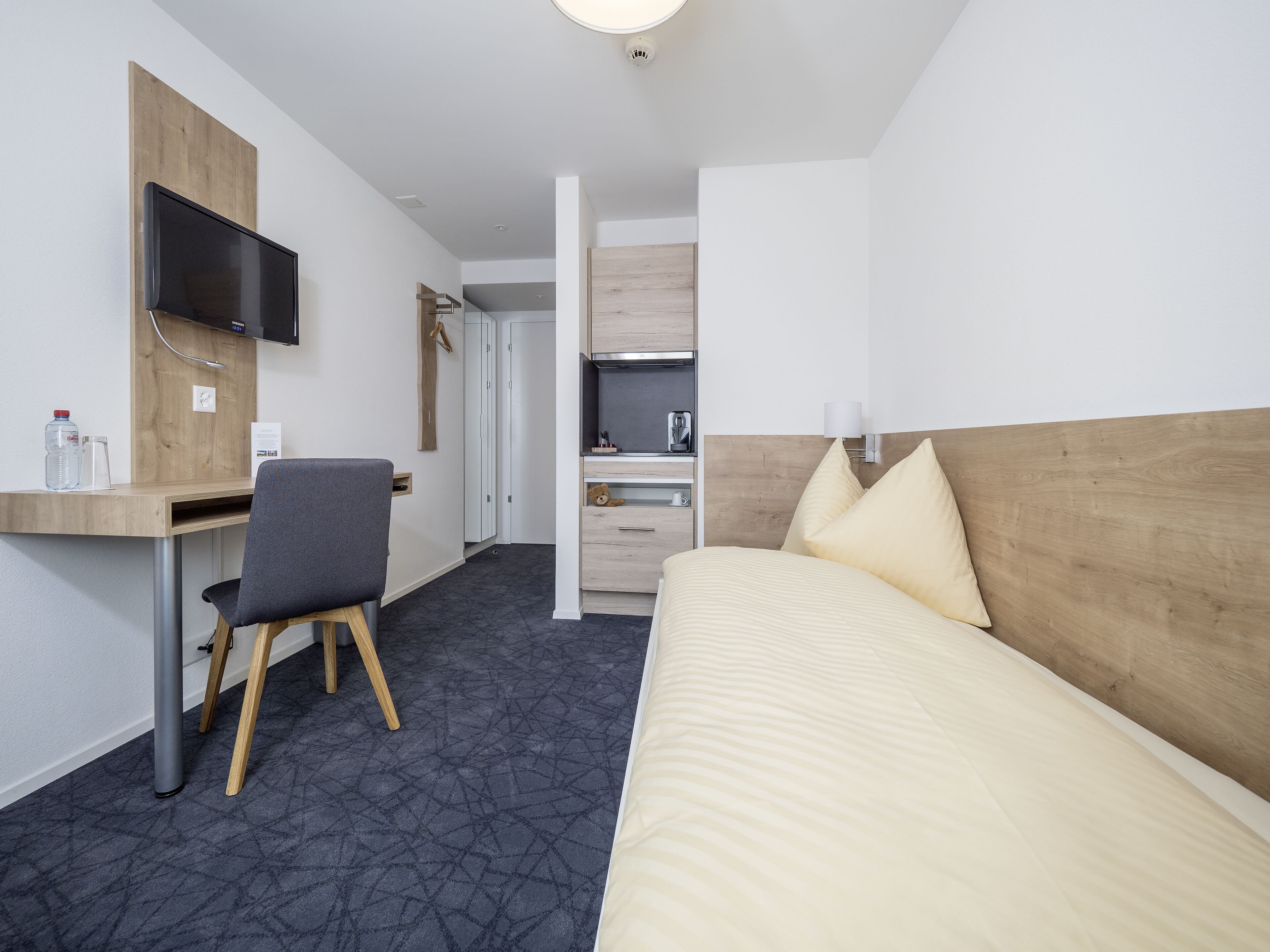 Une chambre d’hôtel comprenant un lit, un bureau avec une chaise, un téléviseur fixé au mur et un coin cuisine.