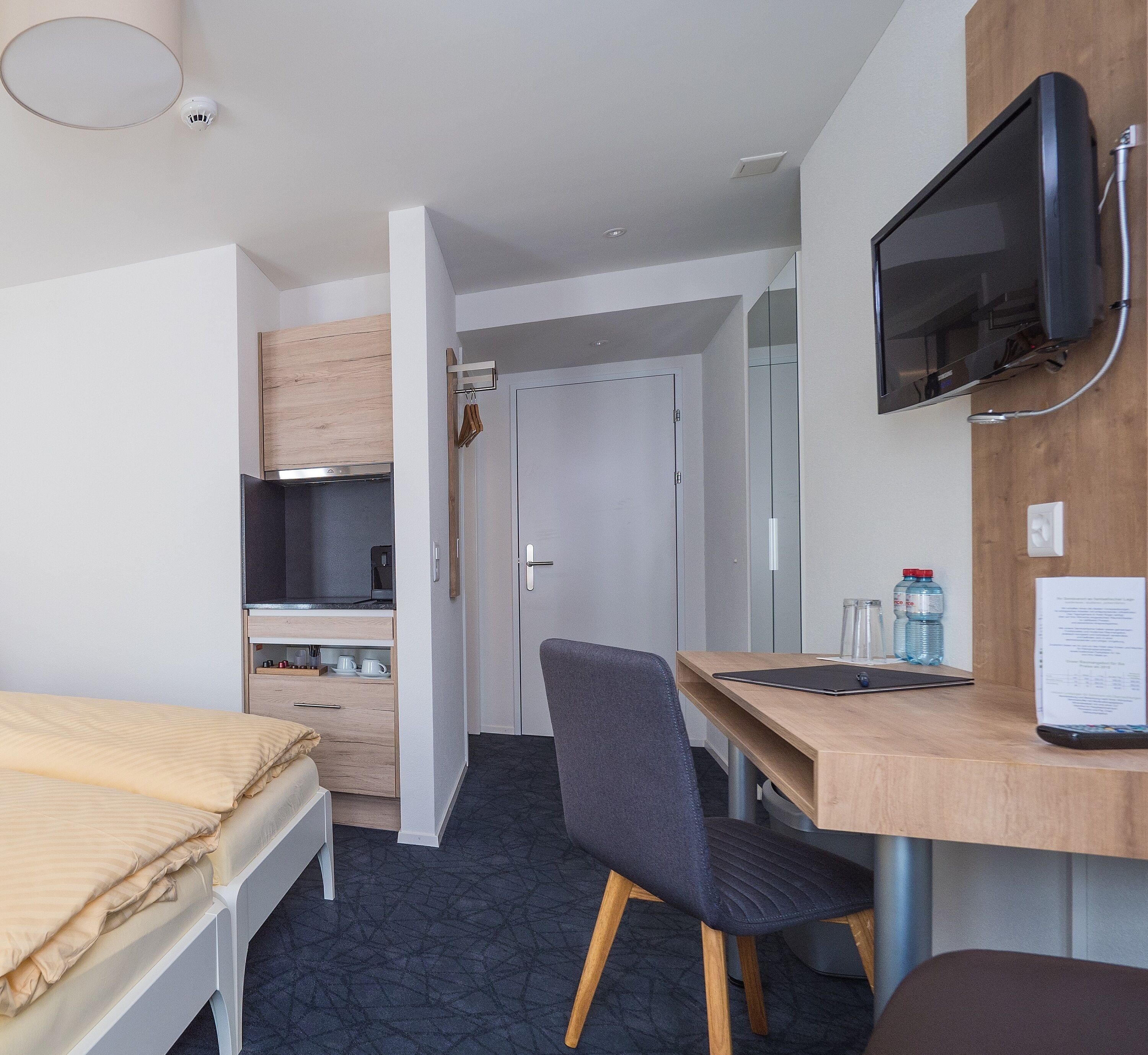 A hotel room with a bed, a desk, a chair, a television, a wardrobe, and a kitchenette.