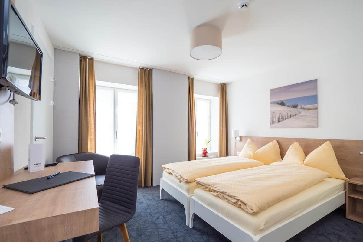 Una camera d'albergo con una scrivania, due letti, una sedia, una televisione e una finestra con tende.