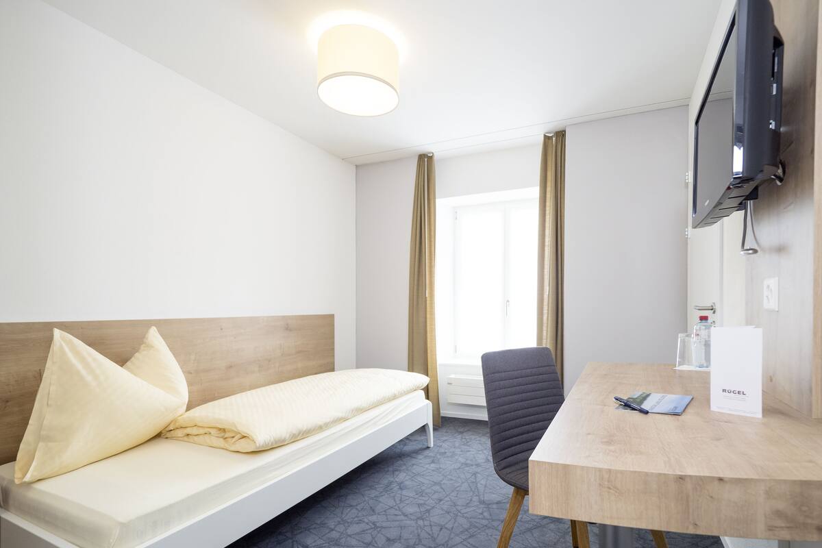 Una camera d'albergo con un letto, una scrivania, una sedia, una televisione e una finestra con tende.