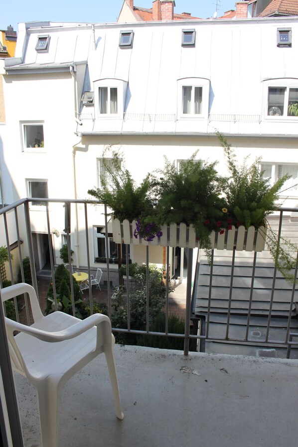 Ein Balkon mit weißem Plastikstuhl, einem hängenden Pflanzgefäß mit Grünpflanzen und Blick auf ein weißes Gebäude mit zahlreichen Fenstern.