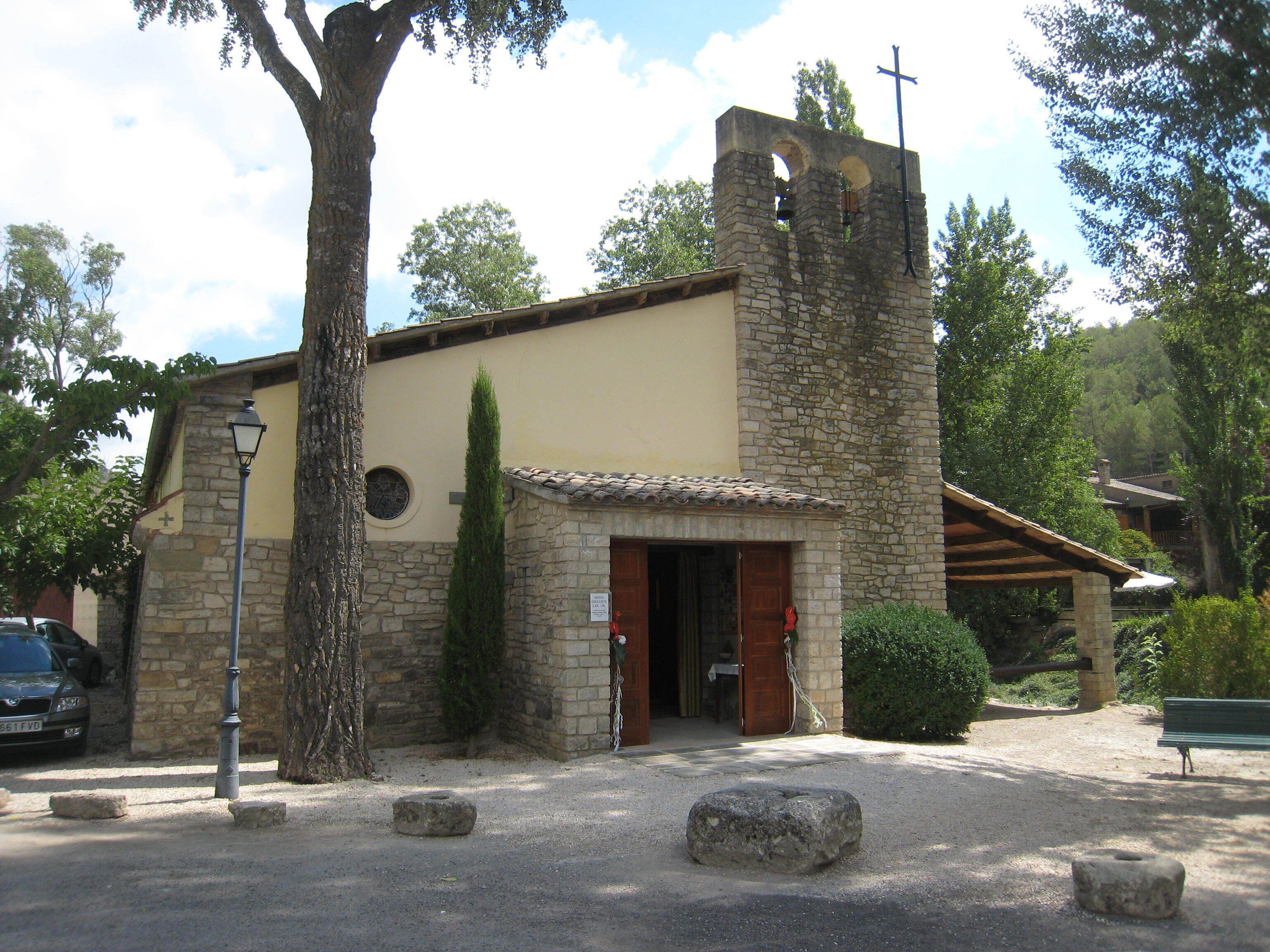 Una iglesia de piedra con un campanario, un árbol grande y un banco en primer plano.