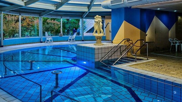 An indoor swimming pool with clear blue water, a central fountain, and a tiled floor. - Hotel Balneari de Vallfogona (Vallfogona de Riucorb)
