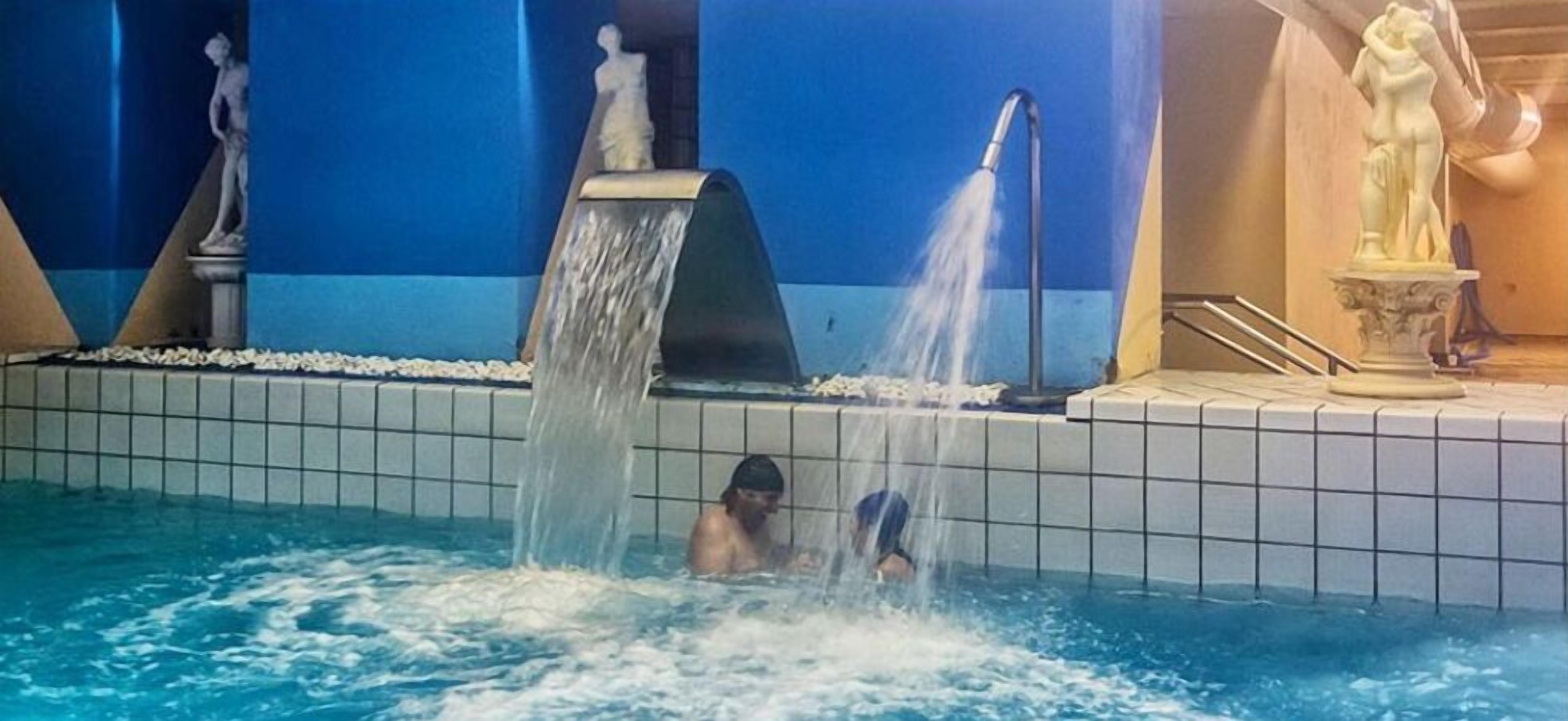 A swimming pool with two people, a water fountain, and a statue.