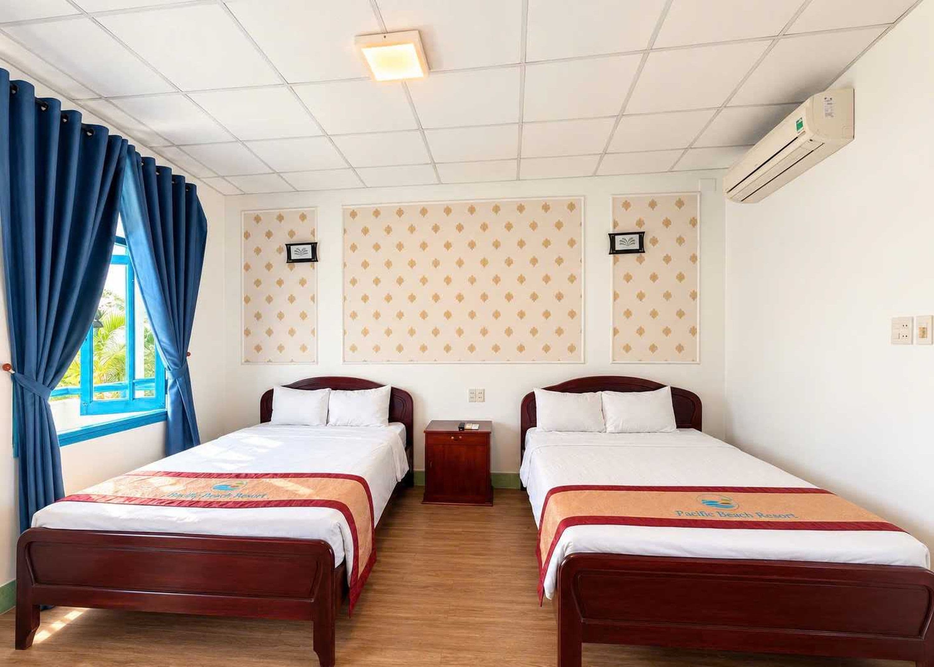 Une chambre d’hôtel avec deux lits, une fenêtre avec des rideaux bleus, un climatiseur fixé au mur et une petite table de chevet.