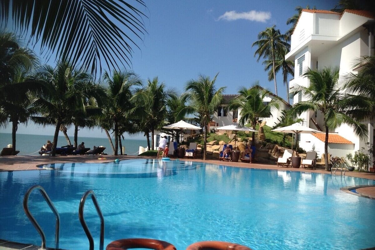 A swimming pool with lounge chairs and umbrellas, surrounded by palm trees and a building with a tiled roof.