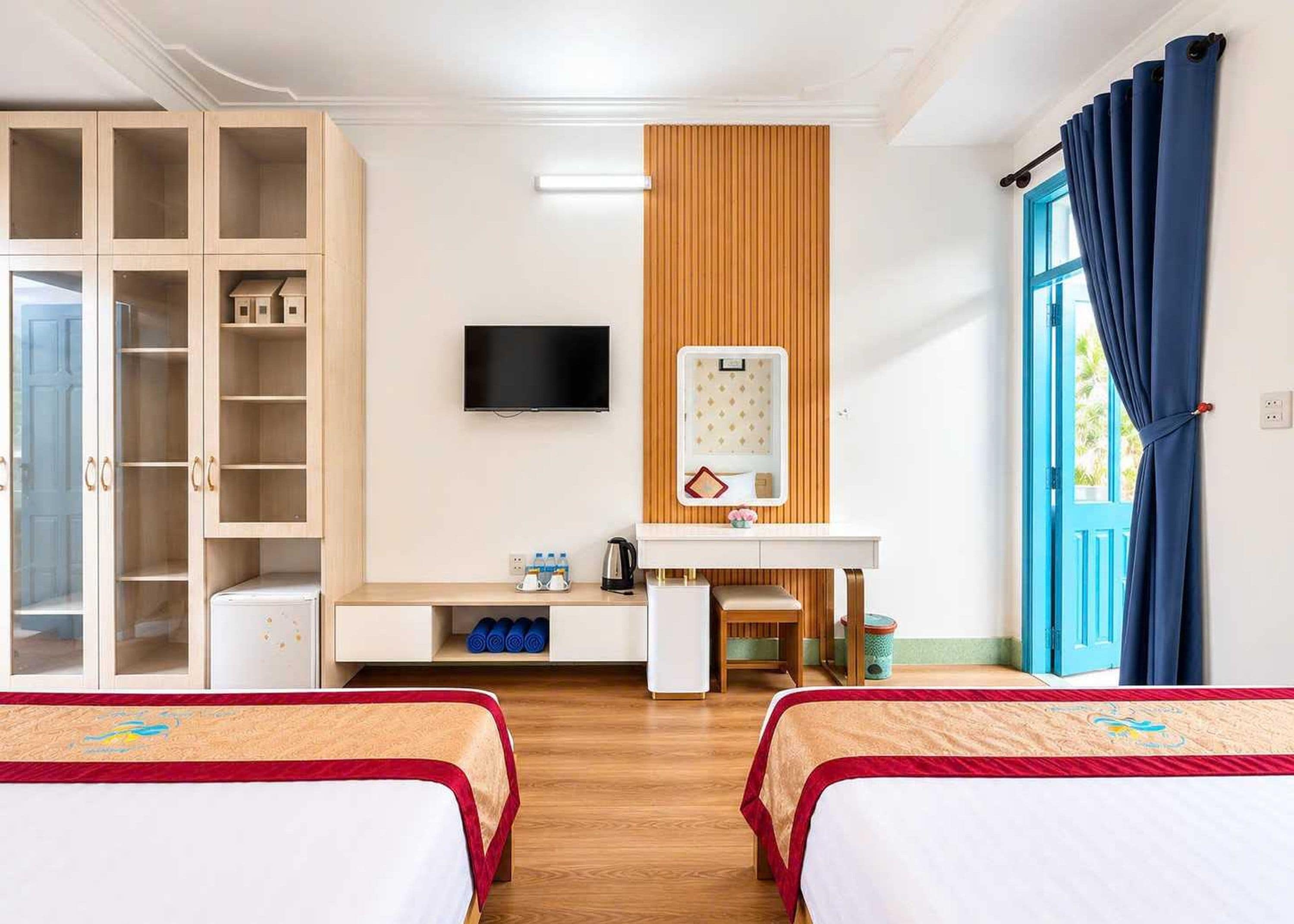 A hotel room with two beds, a wardrobe, a desk with a mirror, a television, and a window with curtains.
