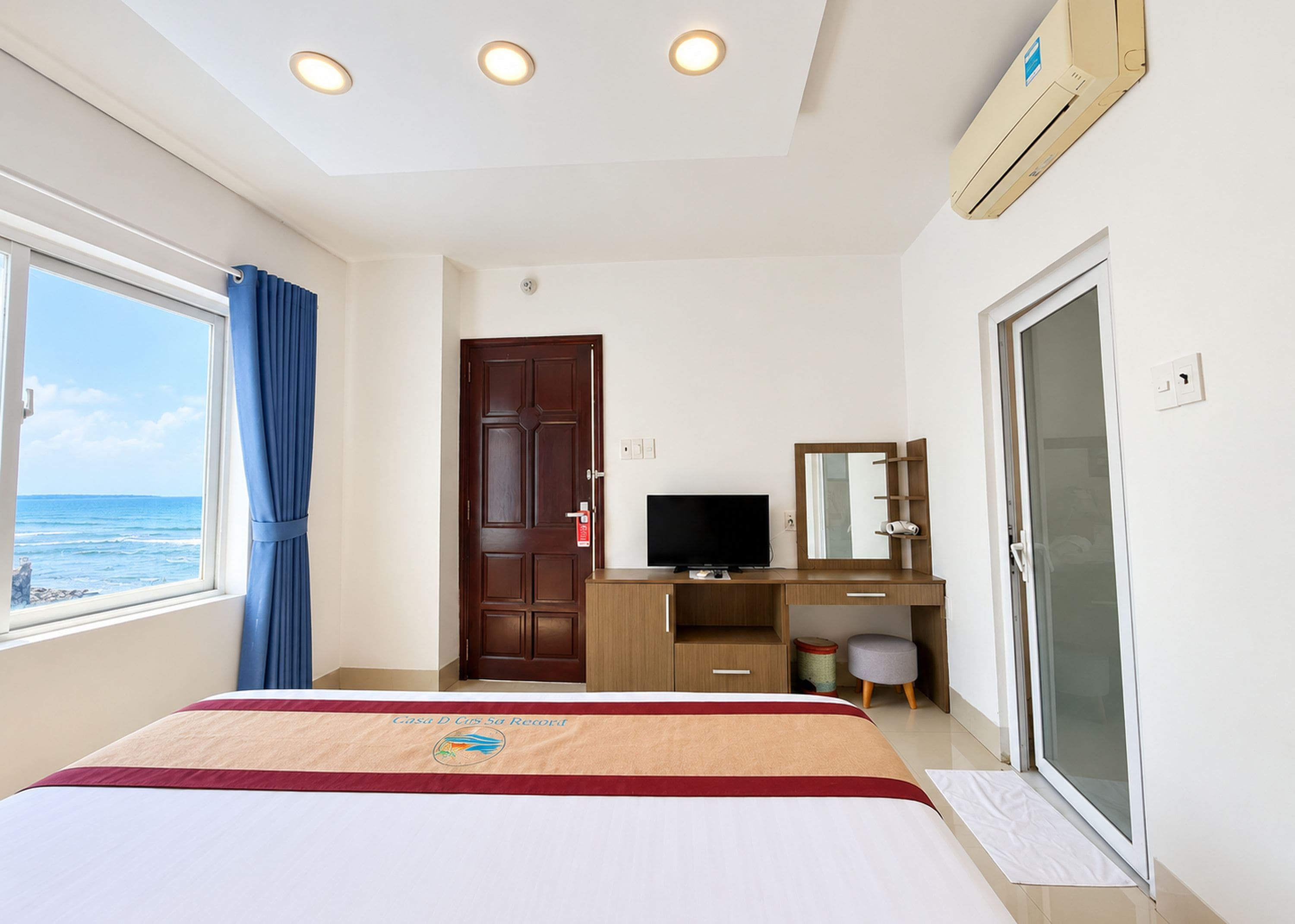A hotel room with a bed, a desk with a television, a window with curtains, and a door.