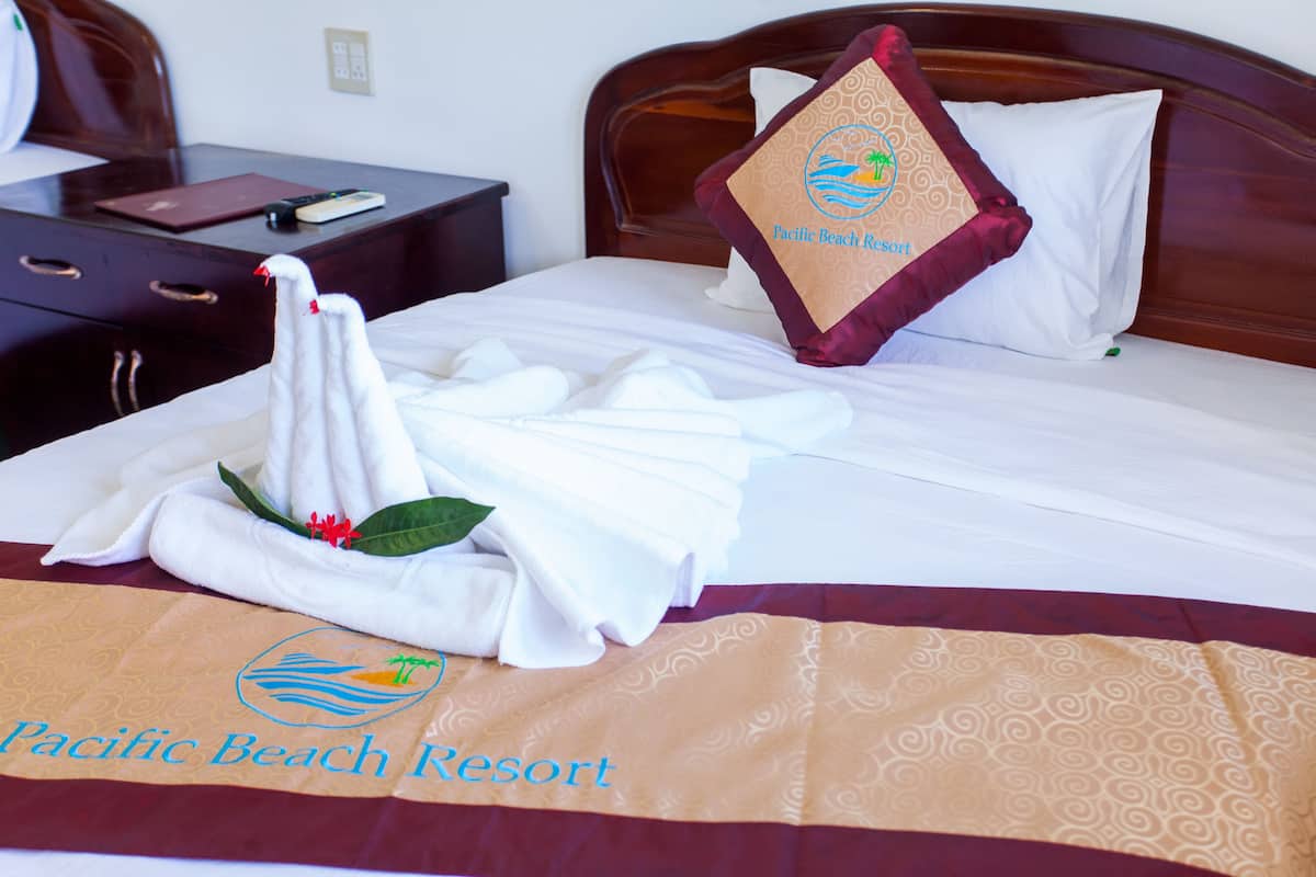 A neatly made bed with a towel and a pillow featuring the Pacific Beach Resort logo.