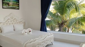 A bedroom with a white ornate bed, white pillows, a nightstand with a lamp, and a view of palm trees through a window. - Pacific Beach Resort (Mui Ne)