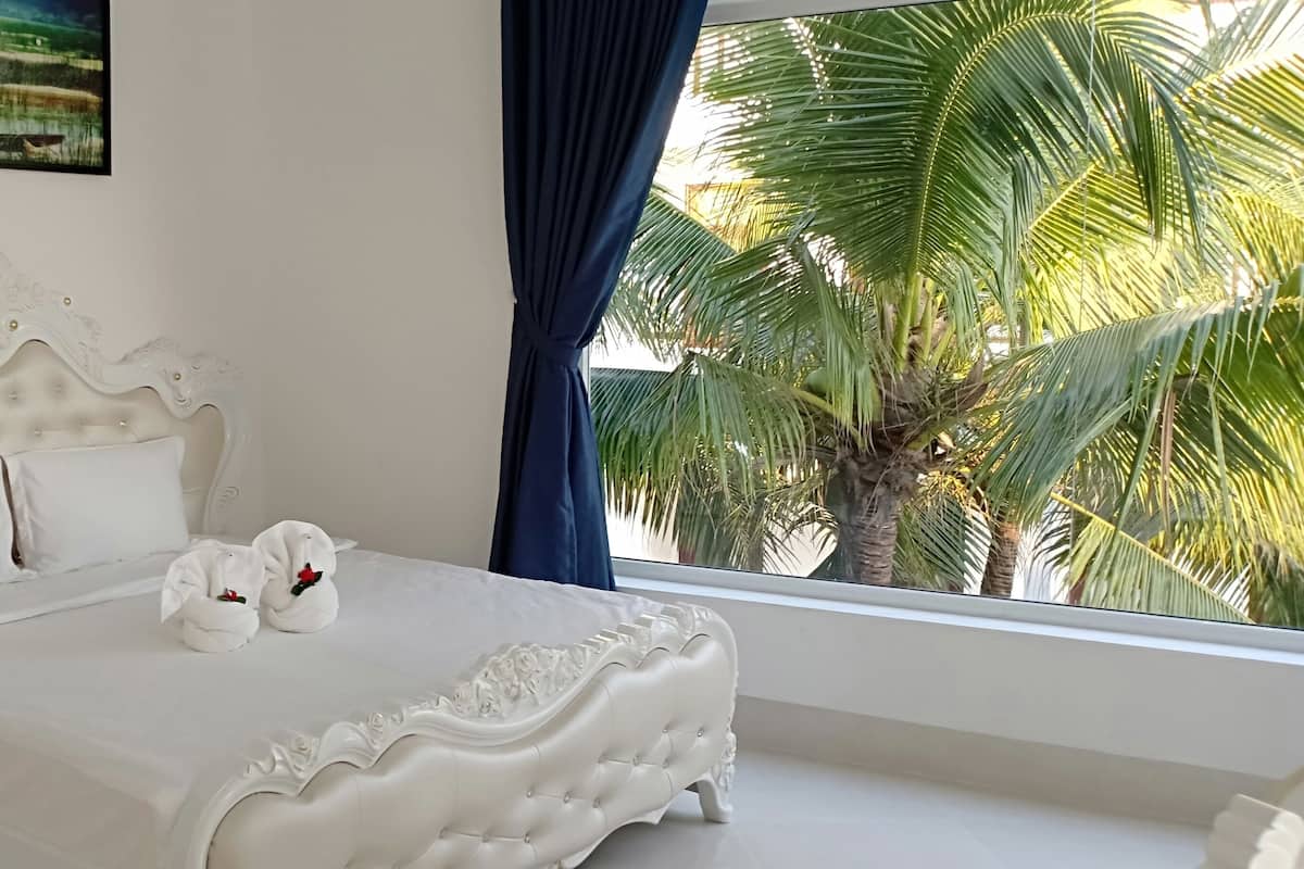 A bedroom with a white ornate bed, white pillows, a nightstand with a lamp, and a view of palm trees through a window.