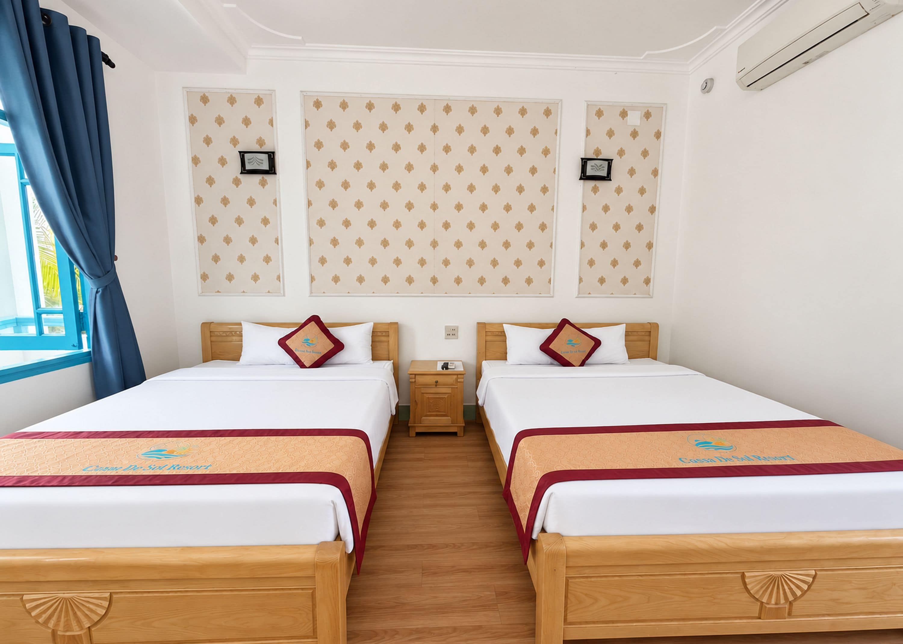 A hotel room with two beds, a wooden headboard, a nightstand, and a window with blue curtains.