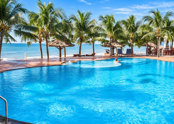 A clear blue pool surrounded by palm trees and lounge chairs, with a view of the ocean. - Sunday Pacific Beach Resort Muine (Mui Ne)