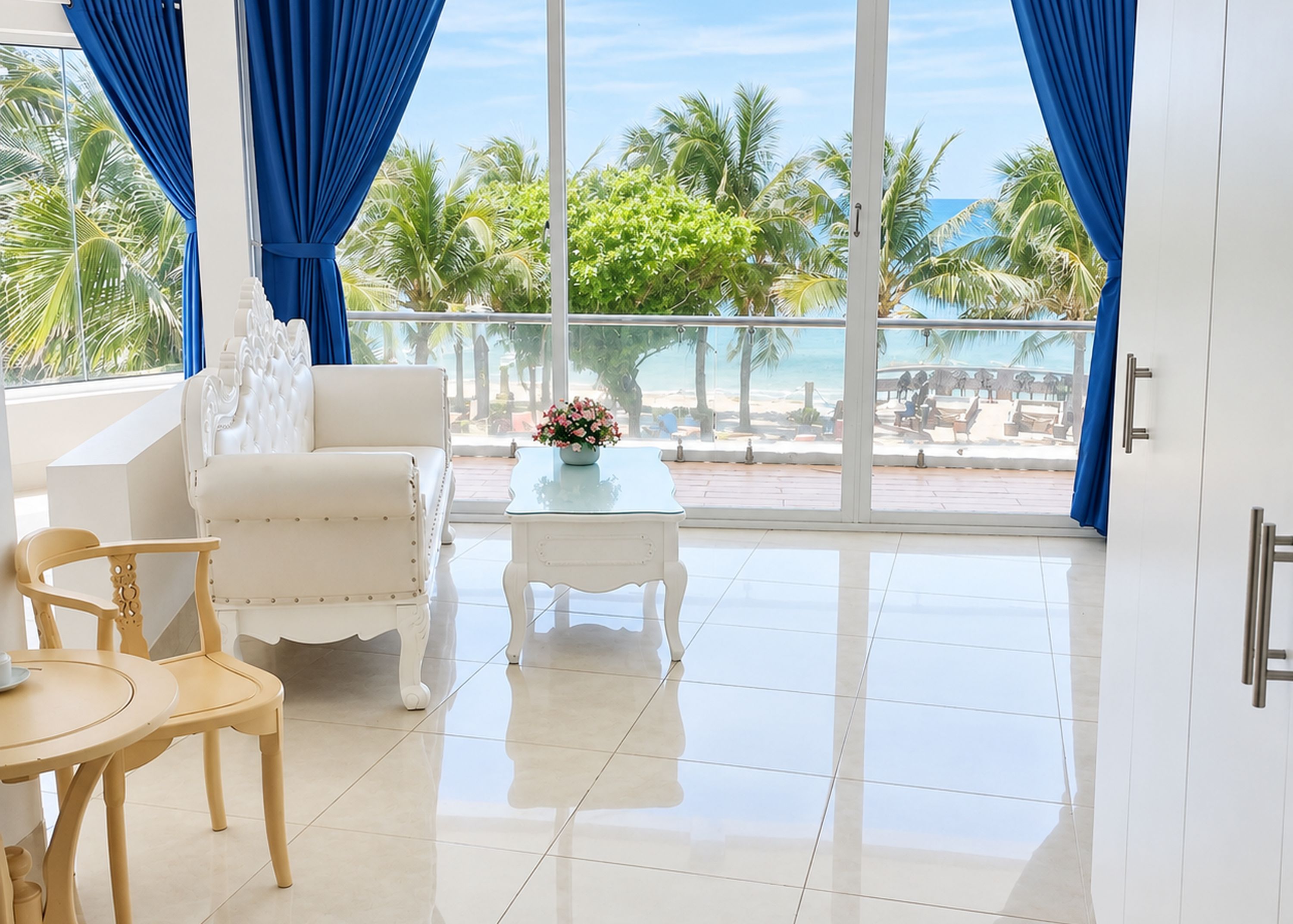 A modern room with a white sofa, a small round table, and a view of a beach and palm trees through large windows.