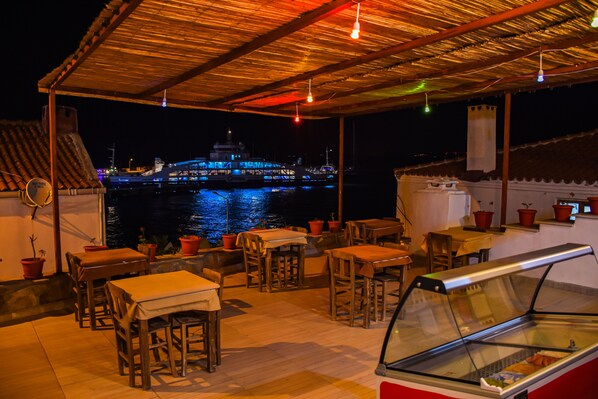 Un espacio al aire libre para comer con mesas y sillas, un expositor de vidrio y vistas a un edificio iluminado al otro lado del agua.