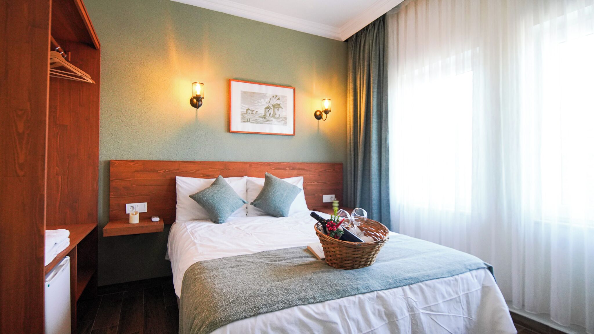 A hotel room with a wooden headboard, a bed with white linens and two pillows, a nightstand with a lamp, a framed picture on the wall, and a window with curtains.