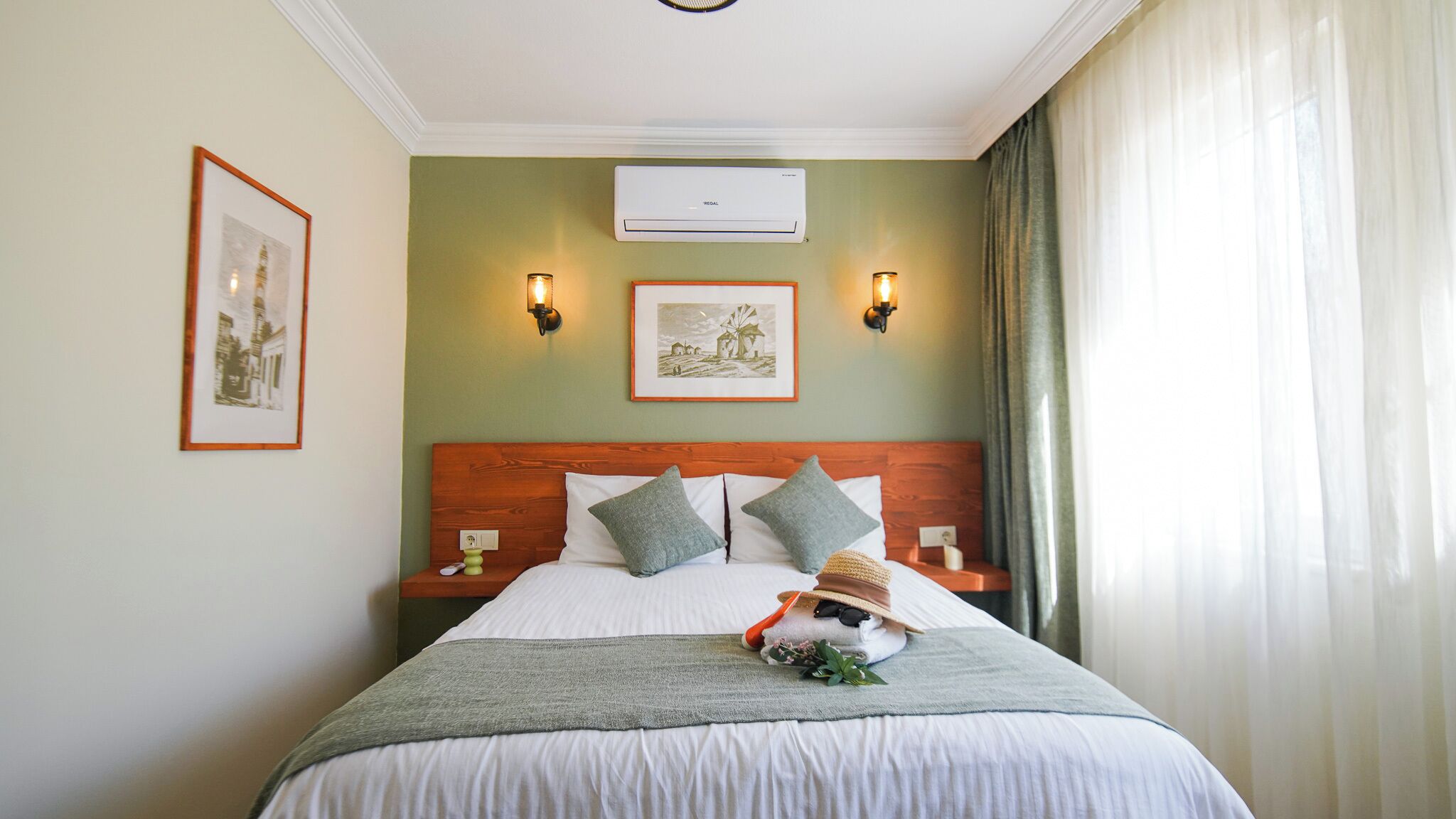 Una habitación de hotel con una cama, dos lámparas de pared, un cuadro enmarcado y un equipo de aire acondicionado.