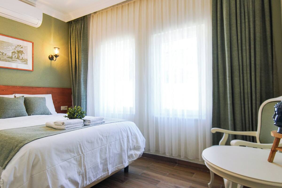 Une chambre d’hôtel avec un lit, une chaise, une table, un tableau et une fenêtre avec des rideaux.