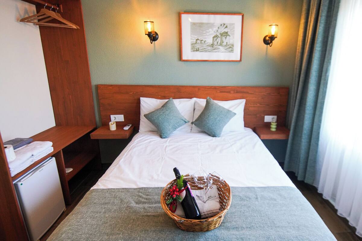Une chambre d’hôtel avec un lit, deux tables de chevet, une armoire en bois, un tableau accroché au mur et un panier contenant une bouteille et des fleurs, posé sur le lit.