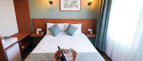 Habitación de hotel con cama, dos mesitas de noche, un armario de madera, un cuadro en la pared y una cesta con una botella y flores sobre la cama.