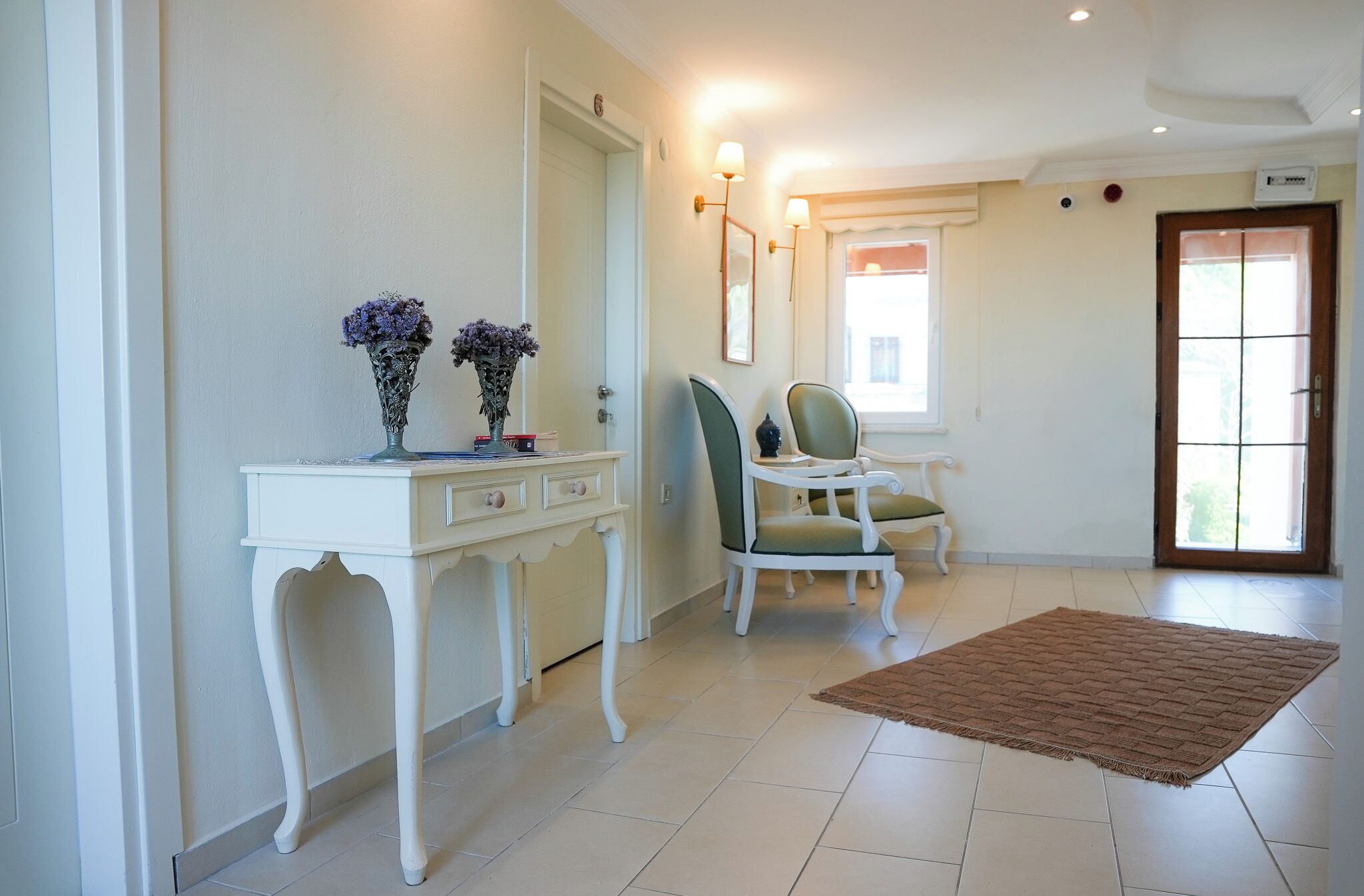 A room with a white desk, two vases with flowers, a green chair, a brown rug, and a door with a window.