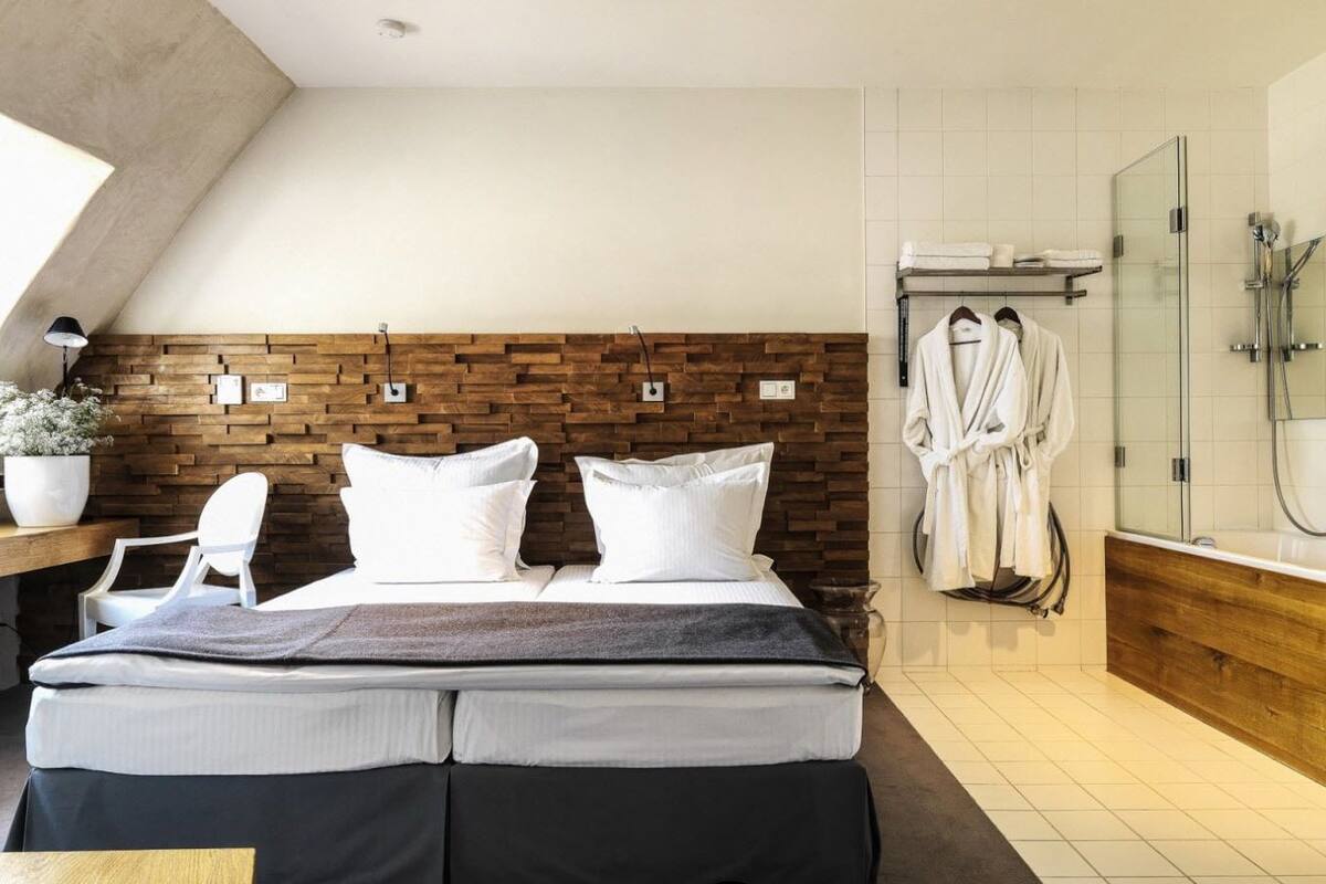 Una habitación de hotel moderna con una cama grande, un cabecero de madera, zona de ducha y baño con bañera y ducha de vidrio.