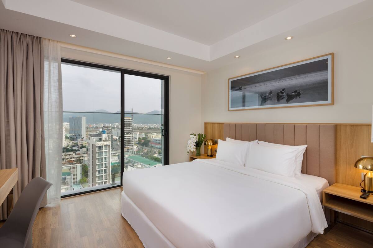 Habitación de hotel con una cama grande, una mesita de noche con lámpara, un cuadro en la pared y vistas a la ciudad a través de un ventanal amplio.